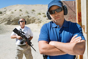A firearms instructor stands with arms folded while another person with a long weapon stands in the background and both are wearing eye and ear protection