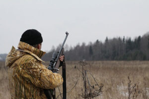 A hunter surveys a pond with a rifle in hand.