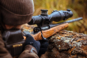 A hunter in the woods rests his rifle with scope on a fallen tree trunk while lining up a shot.