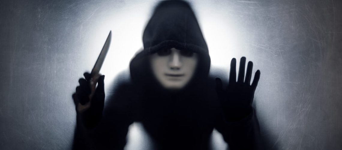 Man in a hood and white mask with knife behind a dusty scratched glass